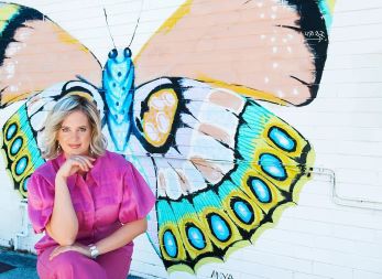 A photo of a blond  lady in pink sitting in front of a brick wall painted with a butterfly