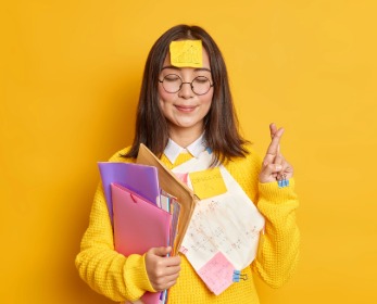 Person standing against a bright yellow background holding folders and papers with sticky notes attached, including one on the forehead, while making a small hand gesture as a reminder cue.