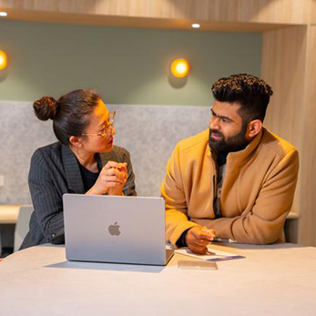 Two people chat, one is an Asian woman wearing glasses, the other is an  man of Indian descent. They sit in front of a laptop.