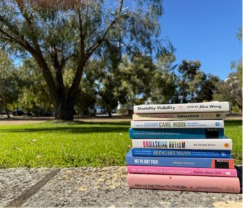 A stack of eight disability-focused books sitting on a stone surface in front of a green lawn and large trees under a clear blue sky.