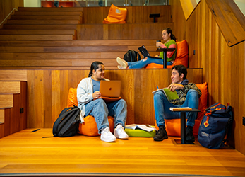 Two students sitting and chatting on a wooden platform, with another student on the stair above. 