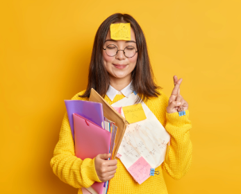 Student holding folders and notes with sticky notes attached to her clothes and forehead, crossing her fingers against a yellow background.
