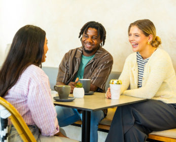 Three students sitting together at a café table, smiling and chatting over drinks.