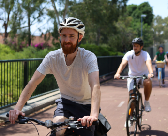Two cyclists riding on a shared path during a guided social bike ride, with trees and greenery in the background.